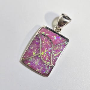 May include: A rectangular pendant with a silver bail. The pendant features a vibrant pink opal stone with iridescent flecks, set within a silver frame. The opal is cut with a geometric design. The jewelry is suitable for a necklace.