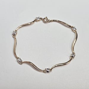 Peut inclure: Un bracelet doré composé d'une série de maillons métalliques lisses et incurvés, reliés par de petits sertis carrés contenant chacun une pierre transparente et brillante. Le bijou est fermé par un fermoir à ressort circulaire.