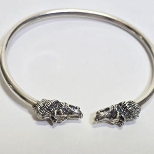 Könnte beinhalten: Ein silberfarbenes Armreif-Armband mit detaillierten Löwenkopf-Terminals. Das Armband hat ein kreisförmiges Design und ist vorne offen. Die Löwenköpfe blicken einander an. Der Schmuck wird auf einer weißen Oberfläche präsentiert.