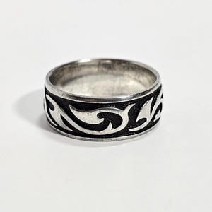 May include: A silver ring with a black, swirling pattern around the band. The ring is circular and sits on a white surface. The design is a repeating, stylized wave or floral motif.