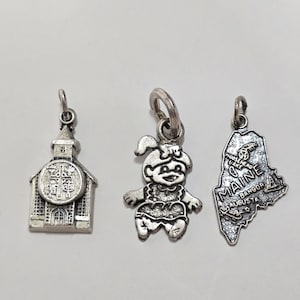 May include: Three silver charm pendants. One is a church, one is a cartoon girl, and one is the state of Maine.