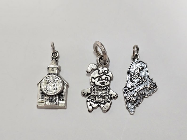 May include: Three silver charm pendants. One is a church, one is a cartoon girl, and one is the state of Maine.