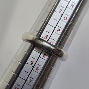 Puede incluir: Un anillo de plata se coloca en un medidor de anillos. El anillo tiene un dise&ntilde;o de banda simple. El medidor de anillos tiene medidas en n&uacute;meros y letras. El anillo est&aacute; en la talla 8.