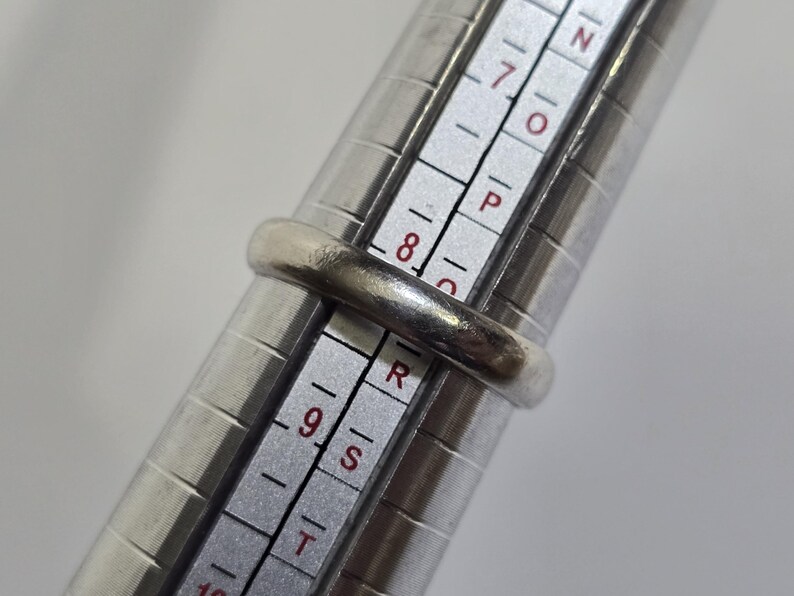 Puede incluir: Un anillo de plata se coloca en un medidor de anillos. El anillo tiene un dise&ntilde;o de banda simple. El medidor de anillos tiene medidas en n&uacute;meros y letras. El anillo est&aacute; en la talla 8.