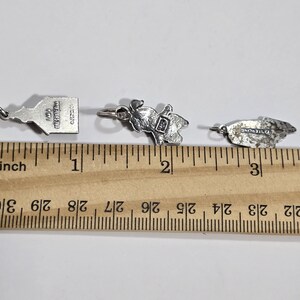 May include: Three silver charms, each about 1 inch long, are shown on a ruler. The charms are a small building with the text "The Metropolitan Museum of Art" on the back, a small figure of a person, and a small leaf.