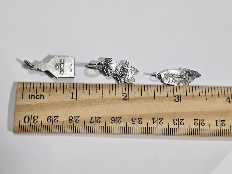 May include: Three silver charms, each about 1 inch long, are shown on a ruler. The charms are a small building with the text "The Metropolitan Museum of Art" on the back, a small figure of a person, and a small leaf.