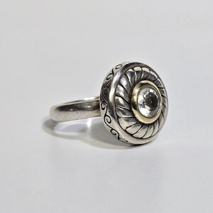 Puede incluir: Anillo de plata con una cara grande y redonda. La cara presenta una piedra transparente central engastada en un bisel dorado, rodeada por un dise&ntilde;o plateado con un borde en forma de cuerda y remolinos decorativos. La banda es lisa y plateada.