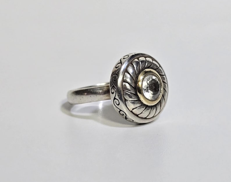 Puede incluir: Anillo de plata con una cara grande y redonda. La cara presenta una piedra transparente central engastada en un bisel dorado, rodeada por un dise&ntilde;o plateado con un borde en forma de cuerda y remolinos decorativos. La banda es lisa y plateada.