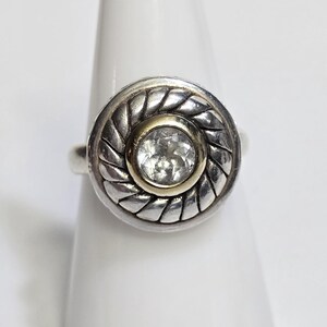 Puede incluir: Un anillo de plata con un centro dorado y una gema redonda y transparente. El anillo tiene un dise&ntilde;o circular texturizado alrededor de la gema. El anillo se muestra sobre una superficie blanca.