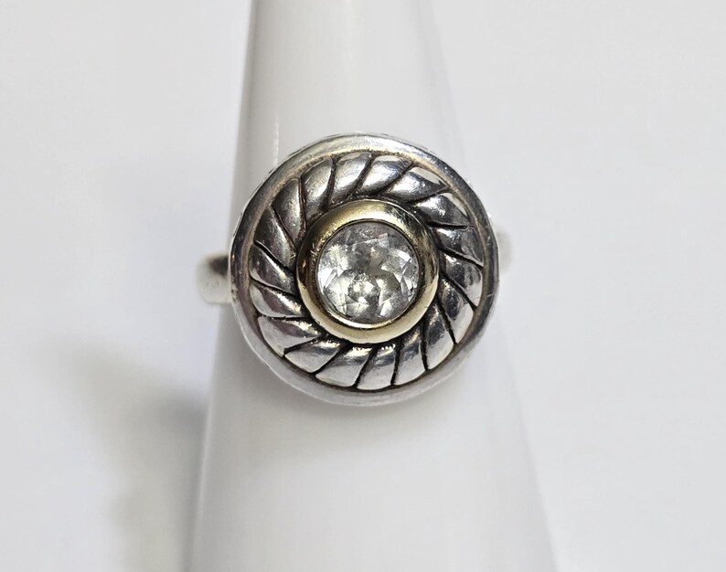Puede incluir: Un anillo de plata con un centro dorado y una gema redonda y transparente. El anillo tiene un dise&ntilde;o circular texturizado alrededor de la gema. El anillo se muestra sobre una superficie blanca.