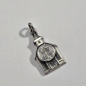 May include: Silver charm in the shape of a church building with a detailed design and a small loop at the top for attaching to a chain or bracelet.