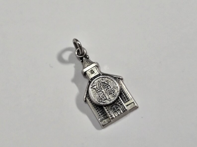 May include: Silver charm in the shape of a church building with a detailed design and a small loop at the top for attaching to a chain or bracelet.