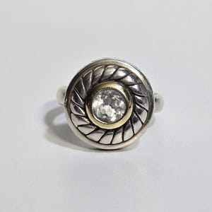 Puede incluir: Un anillo de plata con un dise&ntilde;o circular. El anillo presenta un centro dorado con una piedra clara y facetada. El borde exterior del anillo tiene un patr&oacute;n trenzado.