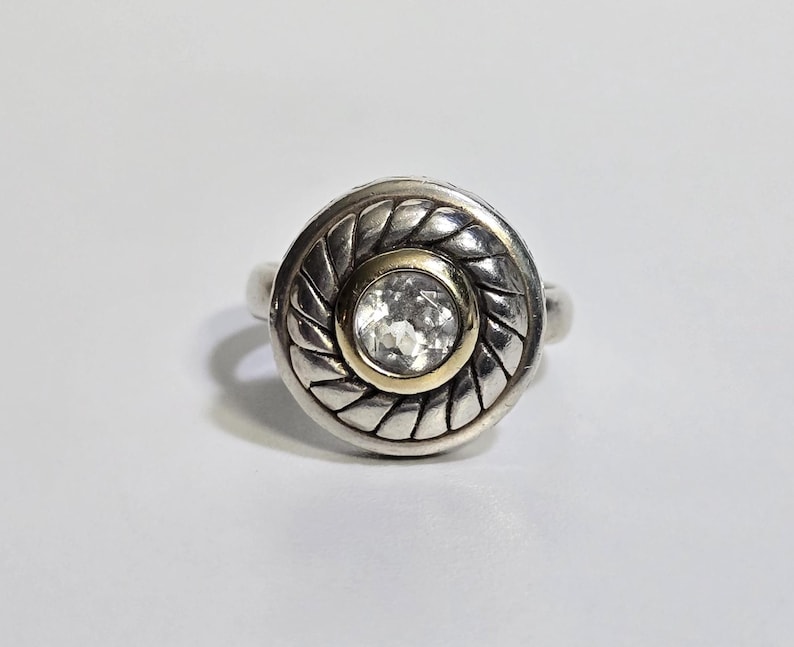 Puede incluir: Un anillo de plata con un dise&ntilde;o circular. El anillo presenta un centro dorado con una piedra clara y facetada. El borde exterior del anillo tiene un patr&oacute;n trenzado.