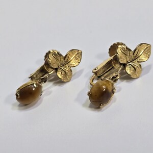 May include: A pair of gold-toned clip-on earrings. Each earring features a leaf design at the top and a brown, oval-shaped stone pendant. The earrings are set against a white background.