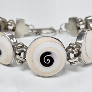 May include: A silver bracelet with a chain link design and round white and black shell accents. The shell accents have a spiral pattern.