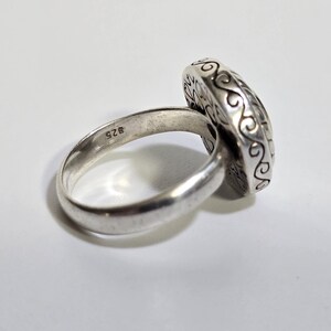 Puede incluir: Un anillo de plata con una cara ovalada y ornamentada. La banda del anillo es lisa y redondeada, con una peque&ntilde;a marca de "925" visible. La cara del anillo presenta un patr&oacute;n detallado y arremolinado en los bordes.
