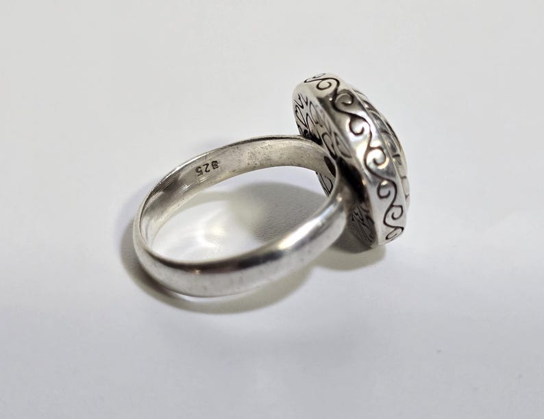 Puede incluir: Un anillo de plata con una cara ovalada y ornamentada. La banda del anillo es lisa y redondeada, con una peque&ntilde;a marca de "925" visible. La cara del anillo presenta un patr&oacute;n detallado y arremolinado en los bordes.