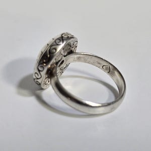 Puede incluir: Un anillo de plata con una cara ovalada y ornamentada. La cara del anillo presenta un dise&ntilde;o detallado con patrones arremolinados y un motivo central. La banda es lisa y pulida, con una peque&ntilde;a marca en forma de coraz&oacute;n. El anillo est&aacute; sobre un fondo blanco.