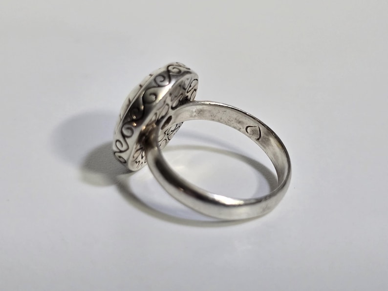Puede incluir: Un anillo de plata con una cara ovalada y ornamentada. La cara del anillo presenta un dise&ntilde;o detallado con patrones arremolinados y un motivo central. La banda es lisa y pulida, con una peque&ntilde;a marca en forma de coraz&oacute;n. El anillo est&aacute; sobre un fondo blanco.