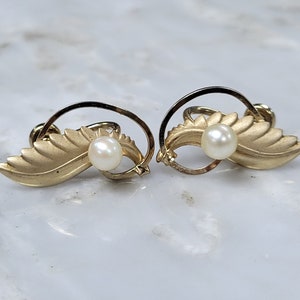May include: A pair of gold-toned leaf-shaped earrings with a pearl accent. The earrings feature a textured leaf design with a small, round pearl in the center. The earrings have a post-back closure.