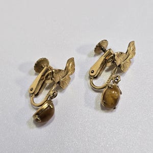May include: A pair of vintage clip-on earrings. The earrings feature a gold-tone metal design with a leaf motif and a teardrop-shaped, brown gemstone. The clip-on mechanism is visible. The earrings are set against a white background.