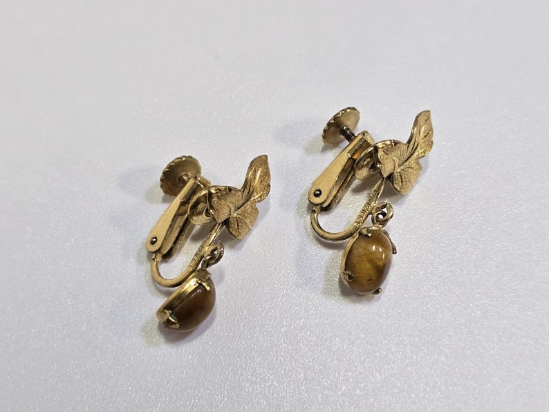 May include: A pair of vintage clip-on earrings. The earrings feature a gold-tone metal design with a leaf motif and a teardrop-shaped, brown gemstone. The clip-on mechanism is visible. The earrings are set against a white background.