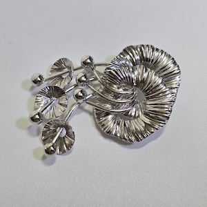 May include: A silver-toned brooch with an abstract design. The brooch features a large, spiraled element and several smaller, circular elements with ball accents. The metal has a reflective, polished finish, creating a modern, artistic look.