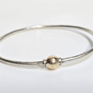 Op de afbeelding: Een zilveren armband met een goudkleurige kraal in het midden. De armband is cirkelvormig en lijkt van metaal te zijn. De gouden kraal vormt een contrast met de zilveren band. Sieraden.