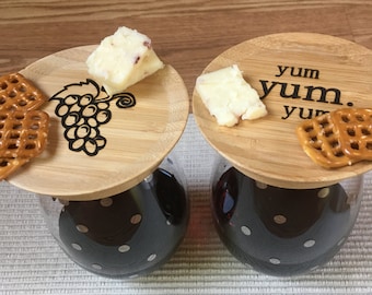 May include: Two stemless wine glasses filled with red wine, each topped with a wooden lid. One lid has a grape design, the other says "yum yum." Each lid holds a piece of cheese and a pretzel.