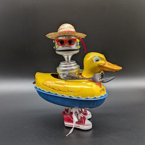 May include: A whimsical sculpture of a yellow duck with a blue base, wearing a straw hat and red sunglasses. The duck has a metal head and is wearing red sneakers. A fork and spoon are placed on the duck's body.