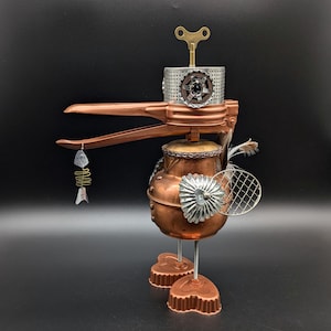 May include: A bird-shaped sculpture constructed from repurposed metal kitchenware, featuring a copper pot body, a long copper beak holding a dangling metal fish charm, a perforated silver head with a brass wind-up key, and heart-shaped feet.