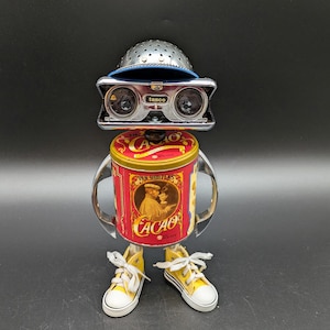 May include: A robot sculpture constructed from a red Van Houten Cacao tin body, chrome binocular eyes labeled Tasco, a silver colander hat, and yellow canvas sneakers for feet. The tin features a vintage illustration of a person drinking cocoa.