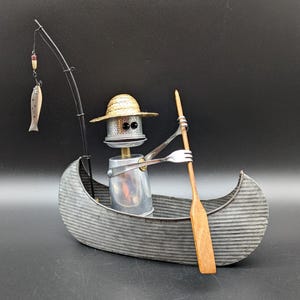 May include: A metal sculpture of a figure in a straw hat sitting in a corrugated gray metal canoe. The figure has a silver metal head and body, holds a wooden paddle, and has a fishing rod with a small metal fish hanging from the line.