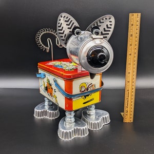May include: A whimsical, handmade metal sculpture of a dog. The head is made from a metal container with a black lens, and the ears are cut from metal. The body is a vintage lunchbox with a colorful design. The legs are made from metal and pie tins.