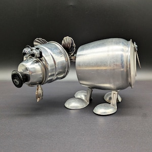 May include: A whimsical, handcrafted sculpture of a pig made from repurposed metal kitchenware. The pig features a round body, a head with two eyes, and four feet. A small fish-shaped charm hangs from the snout. The sculpture is silver in color.