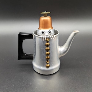 May include: A whimsical, handcrafted teapot with a robot-like design. The aluminum pot features a black handle, a copper-colored top, and brass accents. The robot's face has two black eyes and silver arms. A unique decorative piece.