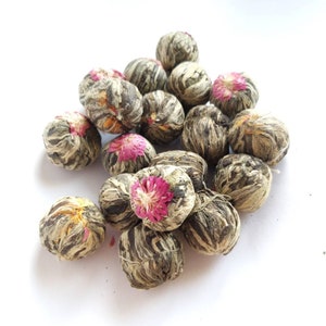 May include: A cluster of flowering tea balls, each encased in dried leaves. Several balls display a bright pink flower at the core. The tea balls are presented on a white background, highlighting their detailed structure and natural hues.