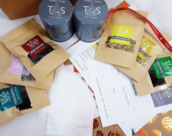 May include: An assortment of tea products, including two gray metal tins, several small kraft paper pouches with tea, and tea filters. The pouches have labels with the text "TSIMSY SUZY" and various tea names. A red ribbon is also visible.