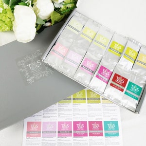 May include: A grey gift box with a clear label and a white box filled with tea bags. The tea bags have colourful labels with the text "TRIMSY SUZY". A sheet of tea descriptions is visible. White flowers are in the background.
