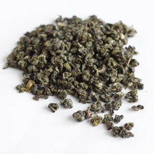 May include: A pile of dried green tea leaves against a white background. The tea leaves are tightly curled and have a slightly glossy appearance. The color is a deep, rich green, suggesting freshness and quality. The image is well-lit, highlighting the texture and detail of the tea.