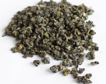 May include: A close-up of a pile of dried green tea leaves on a white surface. The leaves are tightly curled and a deep green colour. The image is well-lit, showing the texture and detail of the tea leaves. The tea appears to be of high quality.