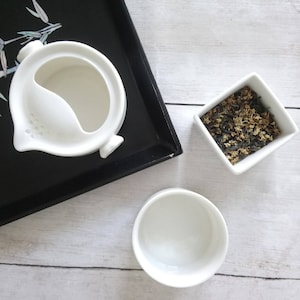 May include: A white ceramic tea set with a teapot, a small bowl, and a teacup. The teapot has a unique spout design. A small square bowl contains dried tea leaves. The set is on a black tray.