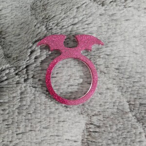 Op de afbeelding: Roze glitterring met een vleermuisvleugelontwerp. De ring is gemaakt van plastic en heeft een diameter van ongeveer 5 cm.