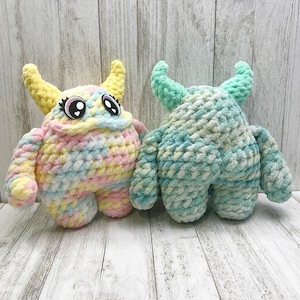 May include: Two handmade crochet monster plush toys. One is pastel-coloured with yellow horns and large eyes. The other is blue and green with curved horns. Both have stubby arms and legs, and are made of soft, chunky yarn.
