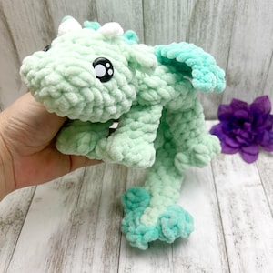 Puede incluir: Un dragón de ganchillo hecho a mano en tonos verde menta y turquesa. El peluche presenta grandes ojos de seguridad negros, cuernos blancos y alas turquesas. El dragón está hecho de hilo suave y texturizado y es una creación caprichosa y achuchable.
