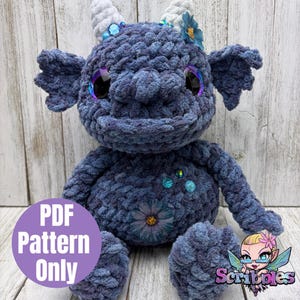 Op de afbeelding: Een handgemaakt, blauw gehaakt wezen met hoorns, oren en grote ogen. Het knuffeldier heeft bloemen en edelsteen versieringen. Een paarse cirkel met de tekst "PDF Pattern Only" staat in de linker benedenhoek.