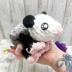 May include: A handmade crochet opossum plush toy. The plush features a white head with black ears, large cartoon eyes, and a pink nose and paws. The body is gray and black. The toy is soft and cuddly, perfect for children or as a decorative item.