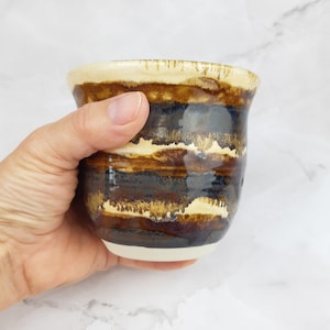 Könnte beinhalten: Eine braune und weiße Keramiktasse mit einem gestreiften Muster. Die Tasse ist handgefertigt und hat eine einzigartige Glasur.
