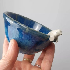 May include: A small blue ceramic bowl with a white ceramic beluga whale figurine attached to the side. The bowl is glazed with a blue and green speckled glaze.